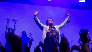A man with arms raised on a stage with band — Foto por Les Taylor na Unsplash